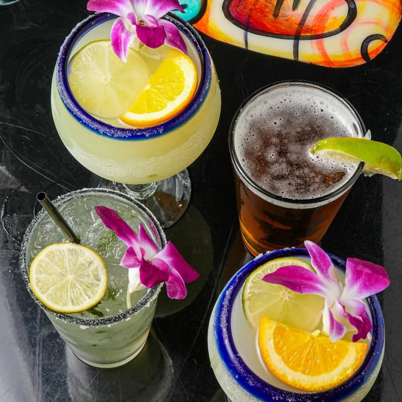 Four colorful Mexican drinks, including margaritas with fruit and orchids, and a beer with lime.