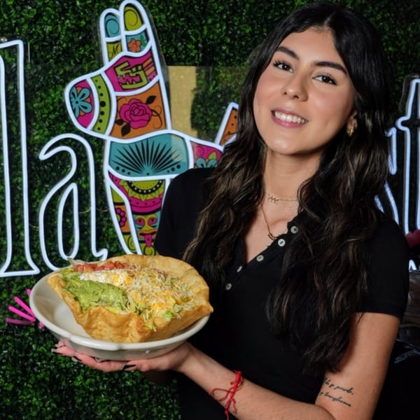 Smiling woman holding a taco salad in a fried tortilla bowl, with a colorful llama sign behind her.