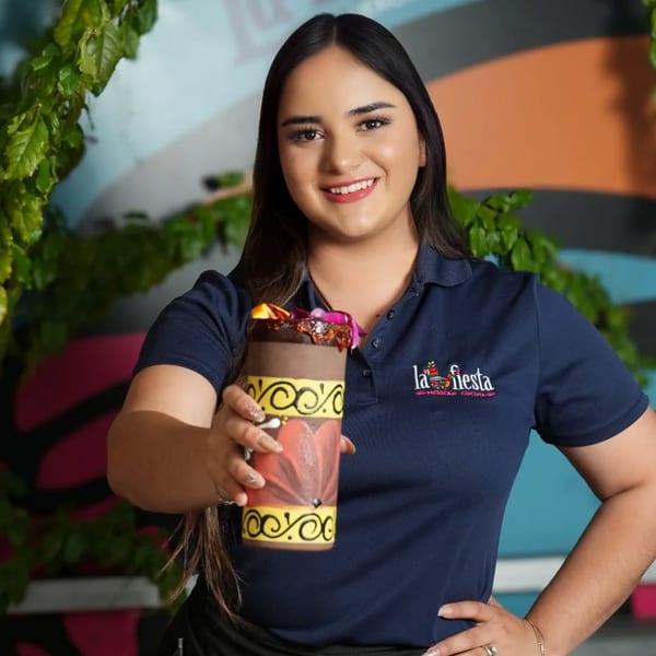 Smiling La Fiesta staff member in a navy polo shirt holding out a colorful Mexican drink.