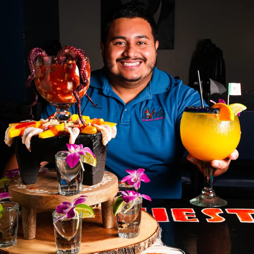 Smiling La Fiesta staff member holding a large orange drink, with a seafood platter and tequila shots.