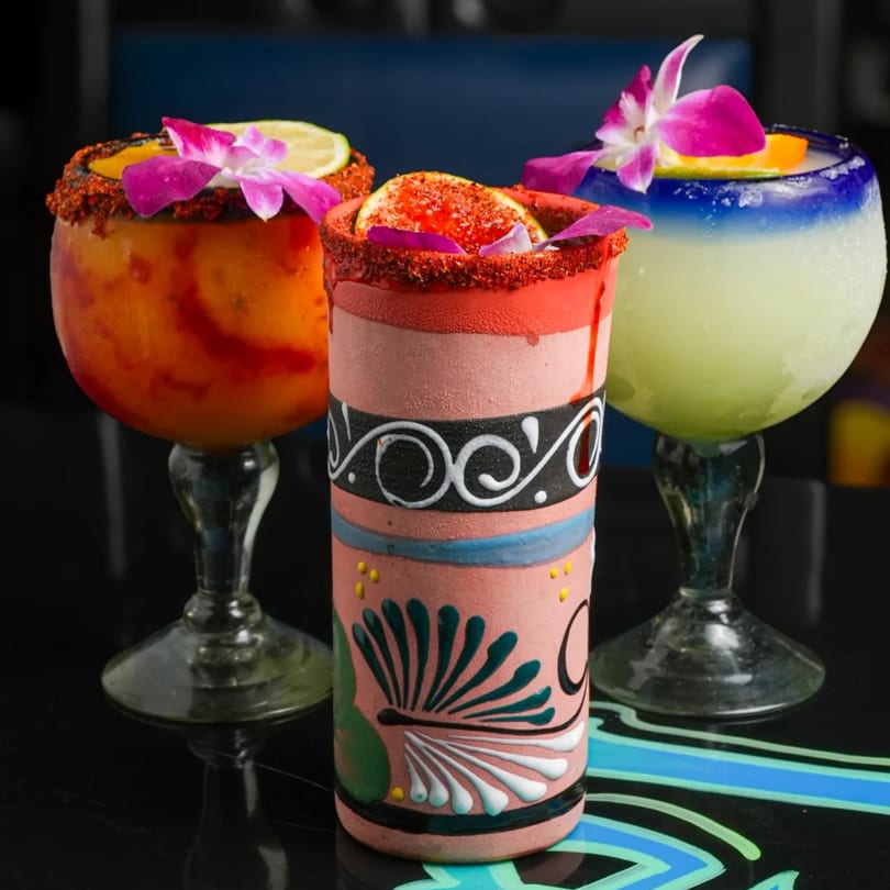 Three vibrant Mexican cocktails, two margaritas in stemmed glasses and one in a decorated ceramic cup.