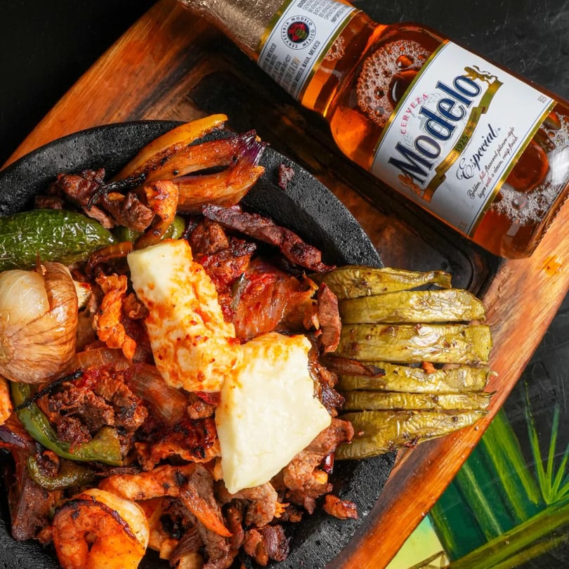 Sizzling Mexican fajitas with grilled meat, vegetables, cheese, and a Modelo Especial beer bottle.